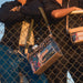 A person in a black pinstripe shirt leans against a chain-link fence holding a small crossbody bag with a leopard-print and graffiti print design. Another person in a denim jacket holds a matching bag on the right.