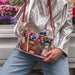 A person wearing a shiny silver jacket and blue jeans carries a clear plastic crossbody bag with a colorful abstract patterned top panel, red strap, and tan leather trim. The bag is shown in an outdoor setting with flowers in the background.