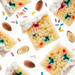 A flat-lay close-up of colorful cake slices topped with white frosting, scattered almonds, and sprinkles on a white background.