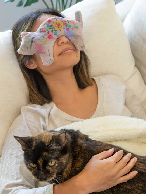 Woman with a floral eye mask holding a cat on a couch