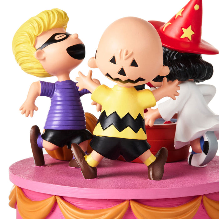 A collectible figurine set of three Peanuts characters dancing on a pink and orange tiered platform against a white background. The characters are dressed in Halloween costumes: a blonde figure in a purple shirt, a bald figure in a yellow and black zigzag shirt, and a figure in a red wizard hat and white robe.