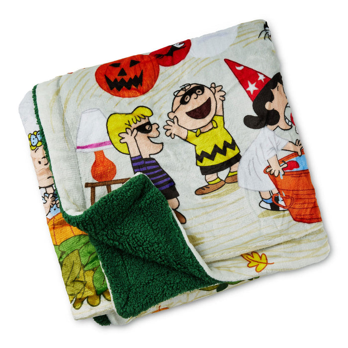 A folded fleece blanket featuring a colorful cartoon print from Peanuts with Halloween characters and pumpkins, showing a dark green sherpa backing.