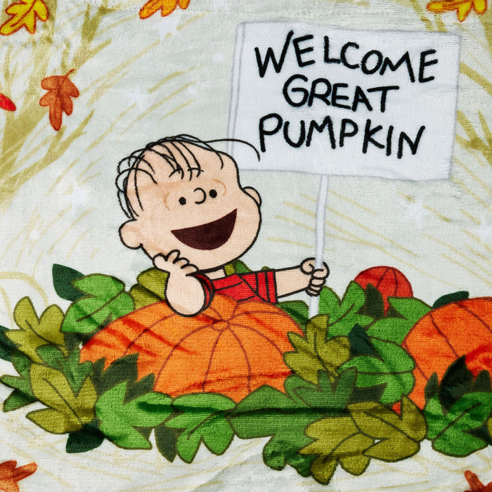 A close-up of a printed textile featuring Charlie Brown holding a sign that reads “WELCOME GREAT PUMPKIN,” surrounded by orange pumpkins and green leaves on a light cream background.