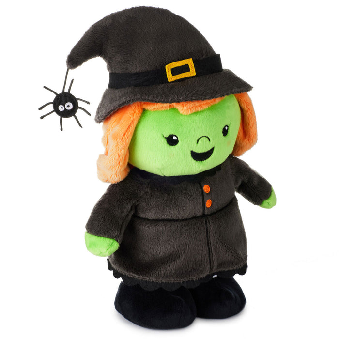 A plush toy dressed as a witch with a green face, orange hair, a black hat and coat, and black shoes, shown on a plain white background with a small black spider accessory near the hat.