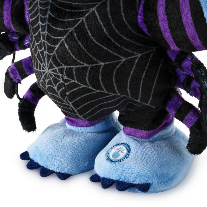 Close-up of a plush monster toy or doll showing the lower body, with a black body printed with a spiderweb pattern, purple striped arms and legs, and light blue feet with dark blue toes. A circular paw-print logo is visible on one foot.