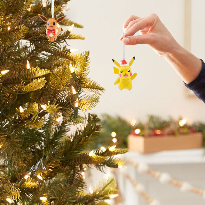 A hand hangs a yellow Pikachu Christmas ornament from a string on a decorated Christmas tree with warm white lights. Another ornament is visible on the tree, and the background shows a softly blurred indoor holiday setting.