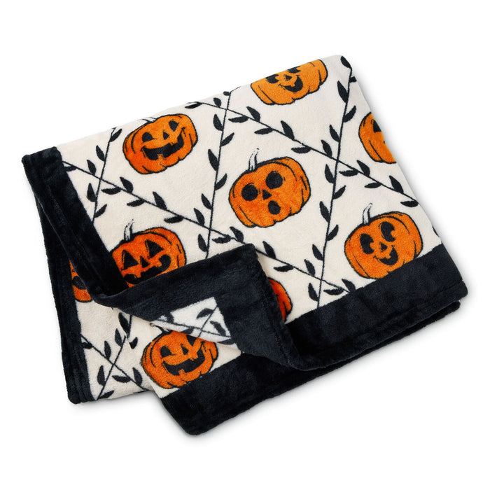 A folded plush blanket with a Halloween theme, featuring orange jack-o'-lantern pumpkins and black vine patterns on an off-white background, finished with a solid black plush border.