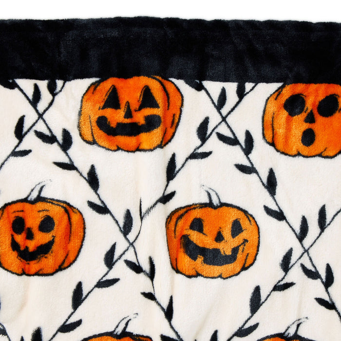 A close-up of a plush blanket featuring a Halloween pattern with orange jack-o'-lantern pumpkins and black leafy vines on an off-white background, finished with a black faux-fur trim.