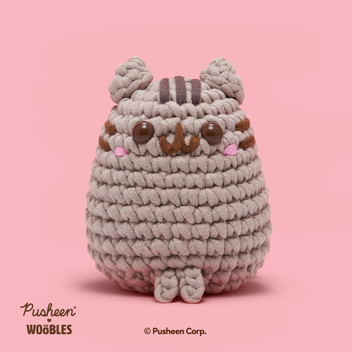 Crocheted plush toy resembling a cat on a pink background, with 'Pusheen' branding.
