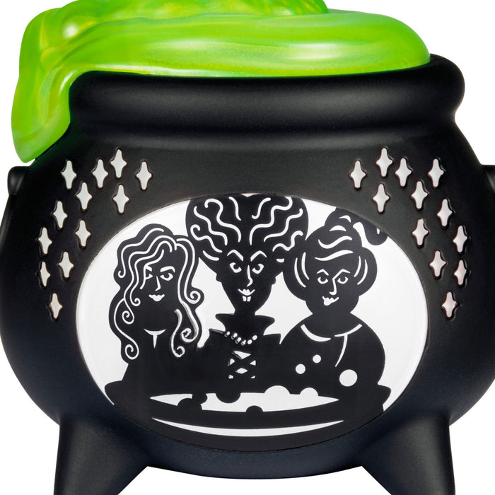 Black cauldron with green lid featuring silhouettes of three women on a white background