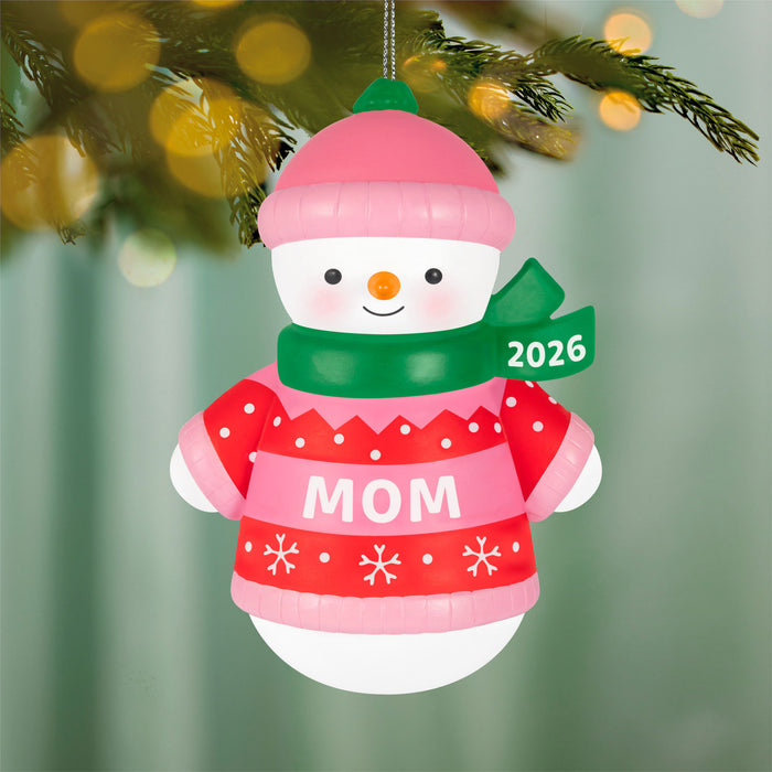 Christmas ornament of a snowman wearing a red sweater with 'MOM' text and a pink hat, hanging on a tree.