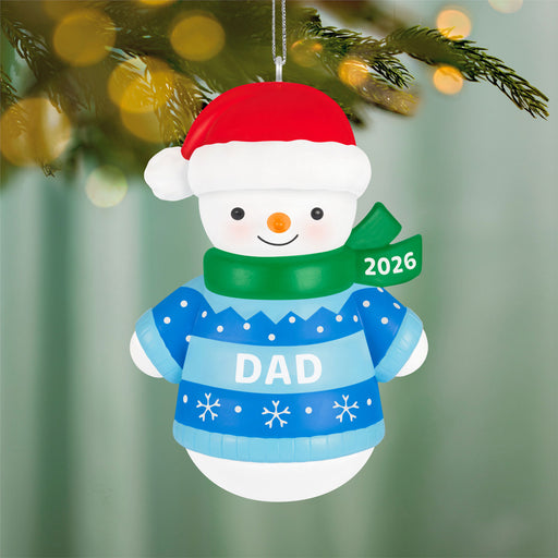 Christmas ornament of a snowman wearing a blue sweater with 'DAD' and a red hat, hanging on a tree.