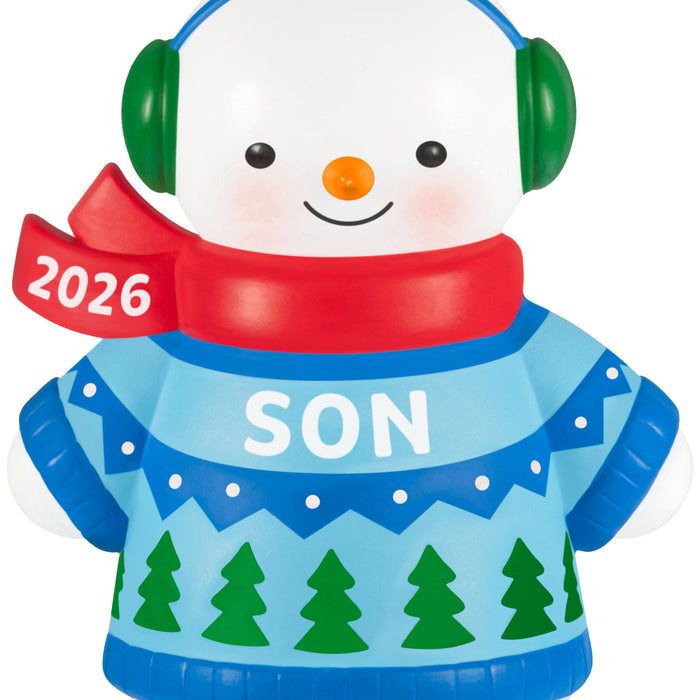 Snowman ornament with 2026 scarf, blue sweater with green trees, and green headphones on a white background.