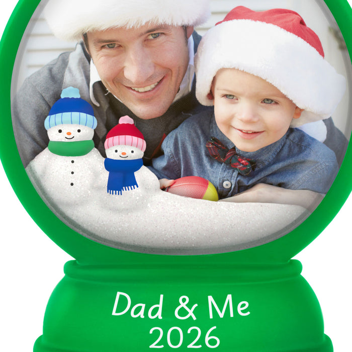 Snow globe ornament with a photo of a man and child wearing Christmas hats, surrounded by snowmen. 'Dad & Me 2026' text on a green base.