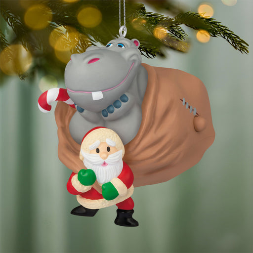 Hippopotamus and Santa Claus Christmas ornament hanging on a tree with blurred lights in the background.