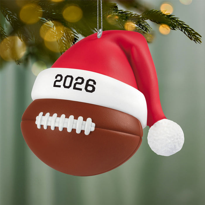 Christmas ornament shaped like a football with a Santa hat and '2026' on it, hanging on a tree.
