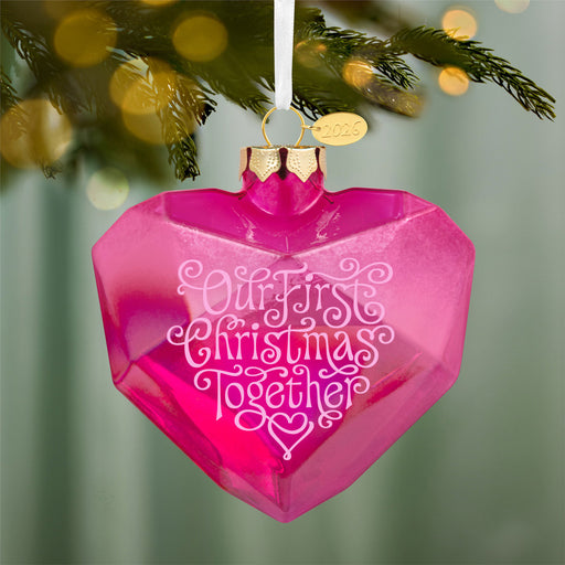 Pink heart-shaped ornament with 'Our First Christmas Together' text on a blurred Christmas tree background.