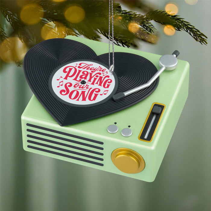 Decorative ornament resembling a record player with 'They're Playing Our Song' text, hanging against a blurred festive background.