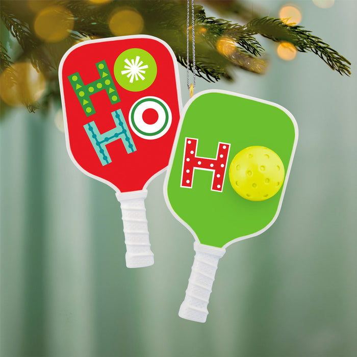 Two Christmas-themed pickleball paddle ornaments with 'HO HO HO' text on a blurred green background.
