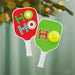 Two Christmas-themed pickleball paddle ornaments with 'HO HO HO' text on a blurred green background.