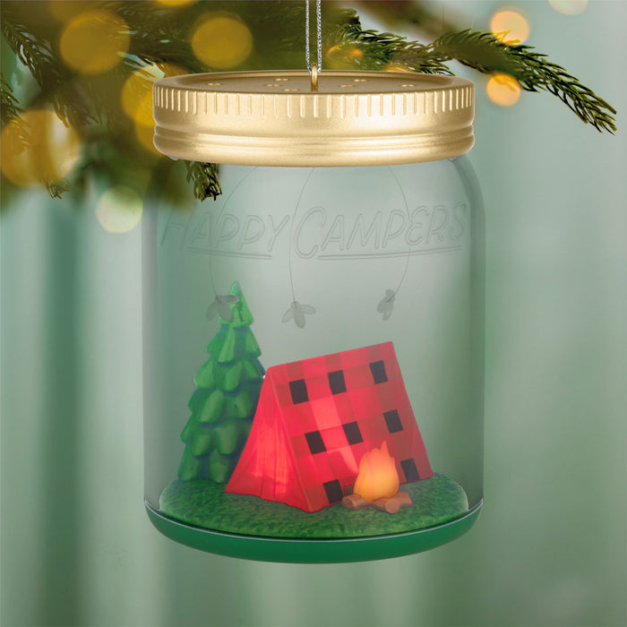 Decorative ornament in the shape of a mason jar with a red tent, green tree, and candle inside, hanging against a blurred green background.