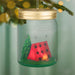 Decorative ornament in the shape of a mason jar with a red tent, green tree, and candle inside, hanging against a blurred green background.