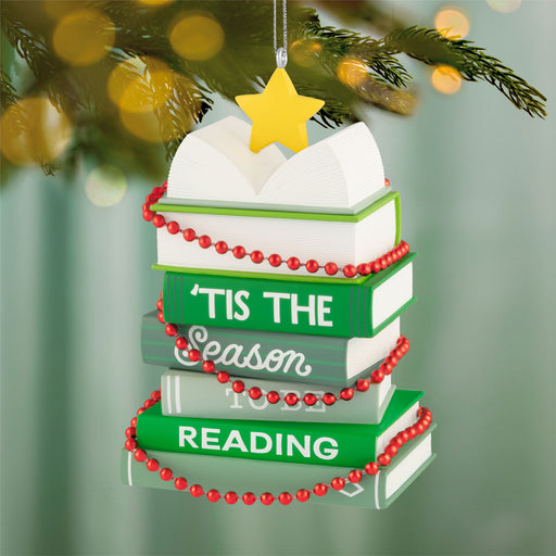 Christmas ornament shaped like a stack of books with 'Tis the Season to be Reading text, decorated with red beads and a yellow star, against a blurred green background.