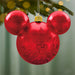 Red ornament shaped like a mouse head with snowflake patterns, hanging on a Christmas tree.