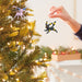Person decorating a Christmas tree with two ornaments, one pink and one black and yellow, against a blurred indoor background.