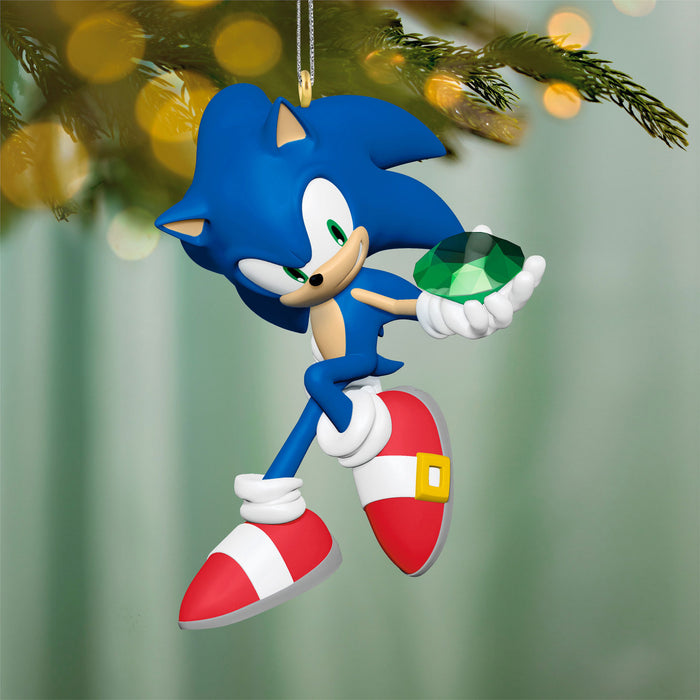 Sonic the Hedgehog Christmas ornament holding a green gem, hanging on a tree with blurred lights in the background.