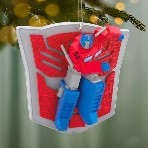 Transformers Christmas ornament with red and blue robot figure on a white shield against a blurred green tree background.