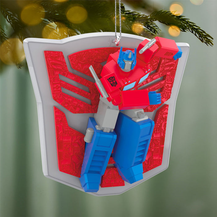 Transformers Christmas ornament with red and blue robot figure on a white shield against a blurred green tree background.