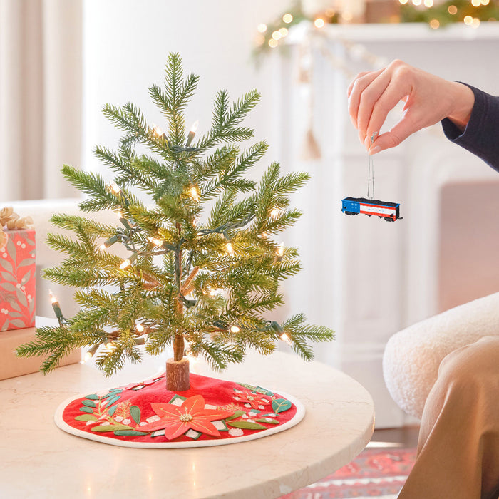 Small decorated Christmas tree with a hand placing a toy train on it in a cozy living room.
