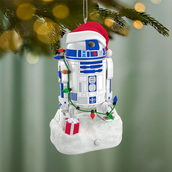 Christmas ornament resembling R2-D2 from Star Wars, decorated with lights and presents, against a blurred green background.