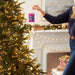 Person decorating a Christmas tree with a colorful ornament in a festive living room.