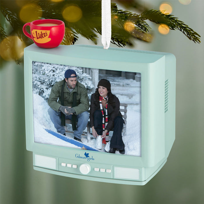 TV-shaped ornament with a scene of two people on skates and a red mug labeled 'Luke's' on a Christmas tree.