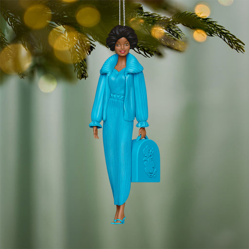 Decorative ornament of a woman in a blue outfit holding a suitcase, hanging from a tree.