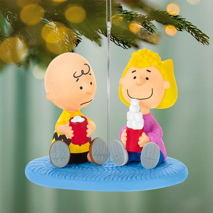 Two Christmas ornaments of a boy and girl sitting on a swing with a blurred Christmas tree background.