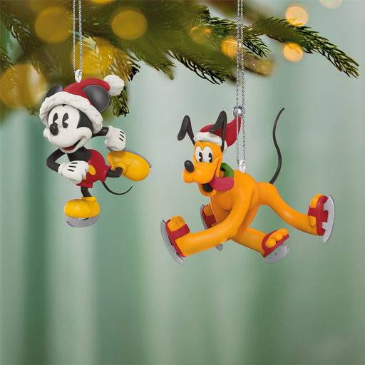Two Christmas ornaments of a mouse and a dog on a tree branch with blurred lights in the background.