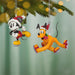 Two Christmas ornaments of a mouse and a dog on a tree branch with blurred lights in the background.