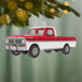 Red and white truck ornament hanging on a Christmas tree branch with blurred lights in the background.