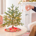 Small decorated Christmas tree on a table with a person's hand holding a ornament in a cozy living room.