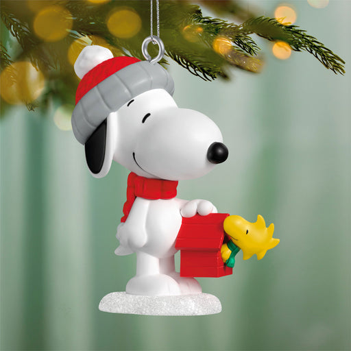 Christmas ornament of a dog wearing a red hat and scarf, holding a yellow bird, on a blurred Christmas tree background.