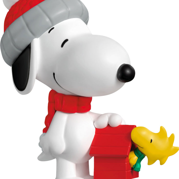 Snoopy figurine with a red hat and scarf, holding a small red house with Woodstock on top.