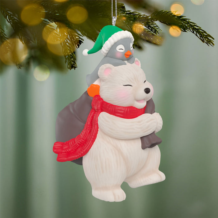 Decorative ornament of a polar bear and penguin hugging, wearing festive hats and scarves, against a blurred Christmas tree background.