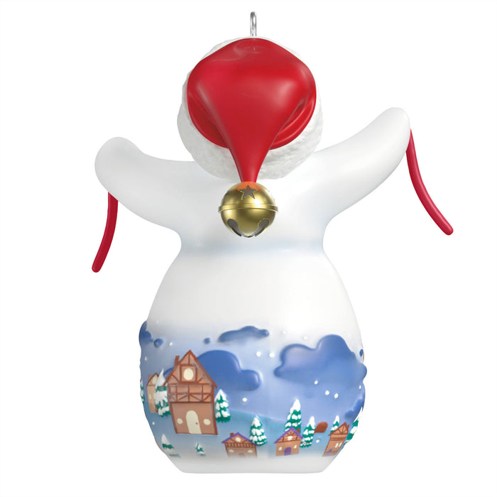 Decorative angel ornament with a red hat and gold bell, featuring a snowy village scene on a white background.
