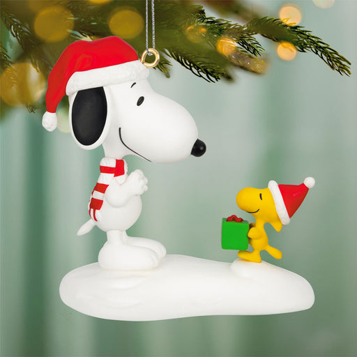 Christmas-themed ornament of Snoopy and Woodstock in Santa hats holding a gift, on a blurred green background.