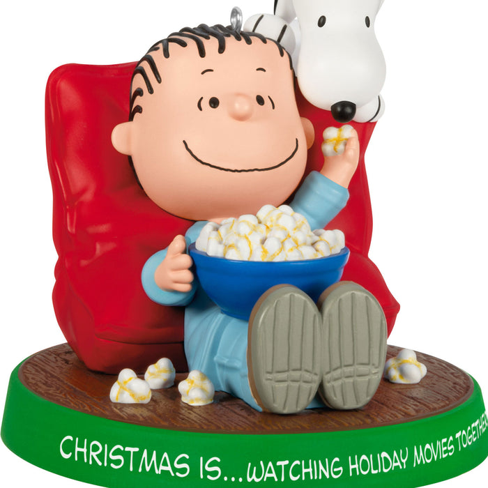 Ornament featuring a character from Peanuts sitting on a red chair with popcorn, next to a dog, on a white background.