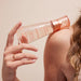 Person applying a bottle of 'Good Chemistry' body mist to their shoulder against a beige background