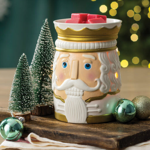 A ceramic nutcracker-shaped candle warmer with a white face, blue eyes, gold trim, and a crown-shaped top holding pink candles, styled on a wooden surface with miniature trees and ornaments.